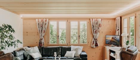 A cozy living room with a sectional sofa, a coffee table, and two armchairs. There are large windows with curtains, a television, and a plant.
