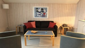 Een woonkamer met een bank, twee fauteuils, een salontafel, een lijst met abstracte kunst, een radio en een lamp.