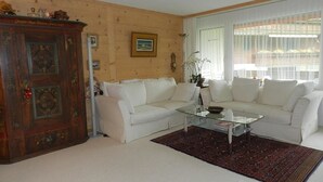 Ein Wohnzimmer mit weißen Sofas, einem Glastisch, einem Holzschrank und einem Fenster mit Vorhängen.