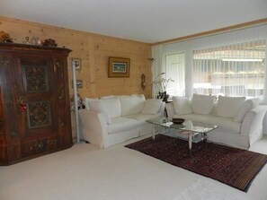 Ein Wohnzimmer mit weißen Sofas, einem Glastisch, einem Holzschrank und einem Fenster mit Vorhängen.