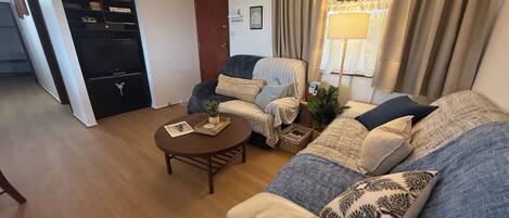 A living room with a sofa, armchair, coffee table, and a small plant. There is a door and a window with curtains.