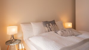 A neatly made bed with white bedding and two pillows, flanked by two bedside lamps on a wooden nightstand. - Fichte (Marija) Appartement/Fewo, Dusche und Bad, WC, 2 Schlafräume by Interhome (Gstaad)