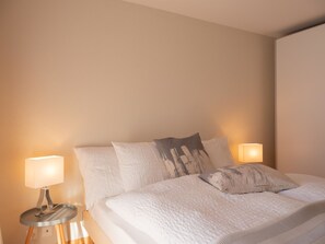 A neatly made bed with white bedding and two pillows, flanked by two bedside lamps on a wooden nightstand. - Fichte (Marija) Appartement/Fewo, Dusche und Bad, WC, 2 Schlafräume by Interhome (Gstaad)