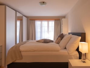 Ein Hotelzimmer mit Bett, Nachttisch, Lampe und einem großen Fenster mit Vorhängen.