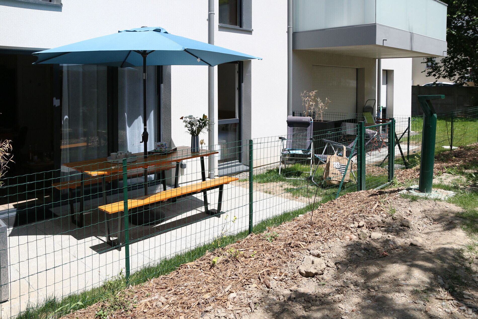 A fenced outdoor area with a table and chairs, a blue umbrella, and a building in the background.