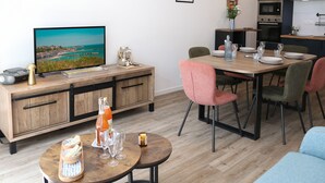 En moderne stue med et træspisebord, to runde borde og en sofa. Et fjernsyn er monteret på væggen, og der er et køkkenområde i baggrunden.