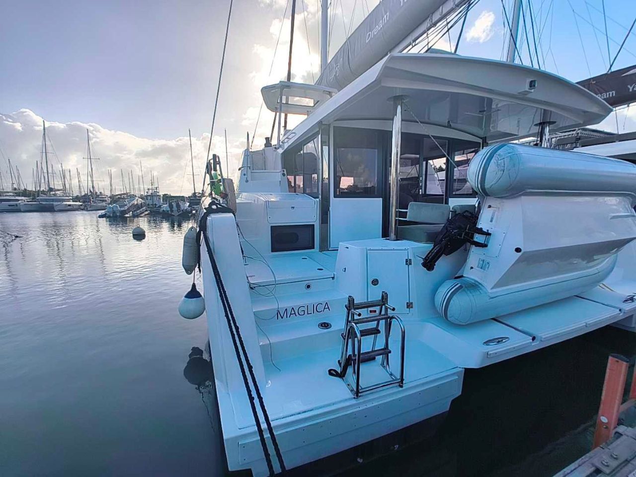 Un yacht blanc nommé MAGLICA était amarré dans une marina, à côté d’autres bateaux. On pouvait y voir une échelle et un treuil sur le pont.