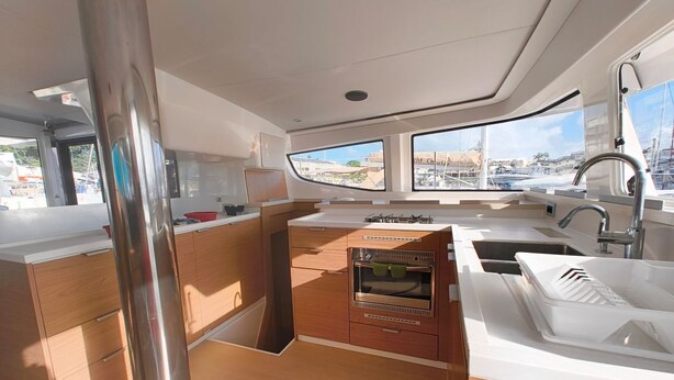 Eine kleine Kombüchenzeile mit Herd, Backofen und Spüle, Holzschränken und Blick auf den Hafen durch ein Fenster.