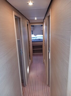 Ein schmaler Korridor mit Holzwandverkleidung und Streifenboden, der zu einem Schlafzimmer mit Bett und Fenster führt.