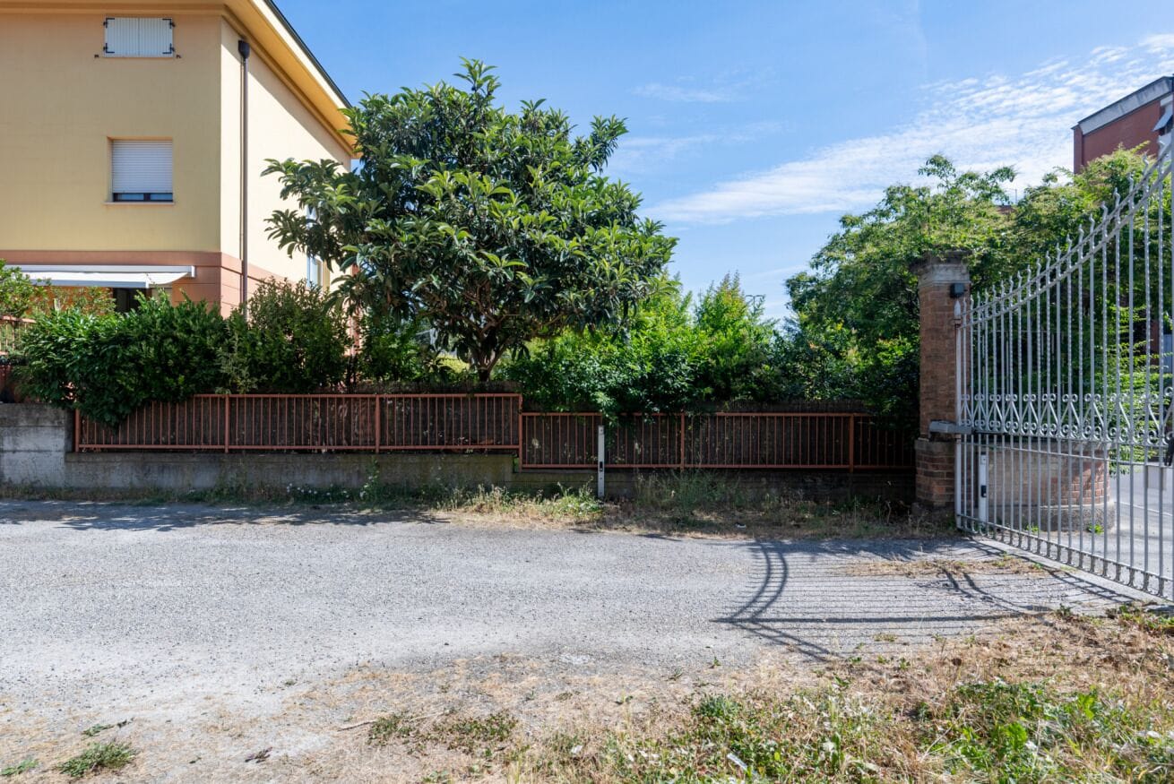 A gated driveway leading to a building with a yellow facade, surrounded by greenery and a metal fence.