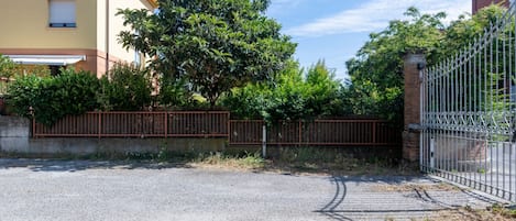 A gated driveway leading to a building with a yellow facade, surrounded by greenery and a metal fence.