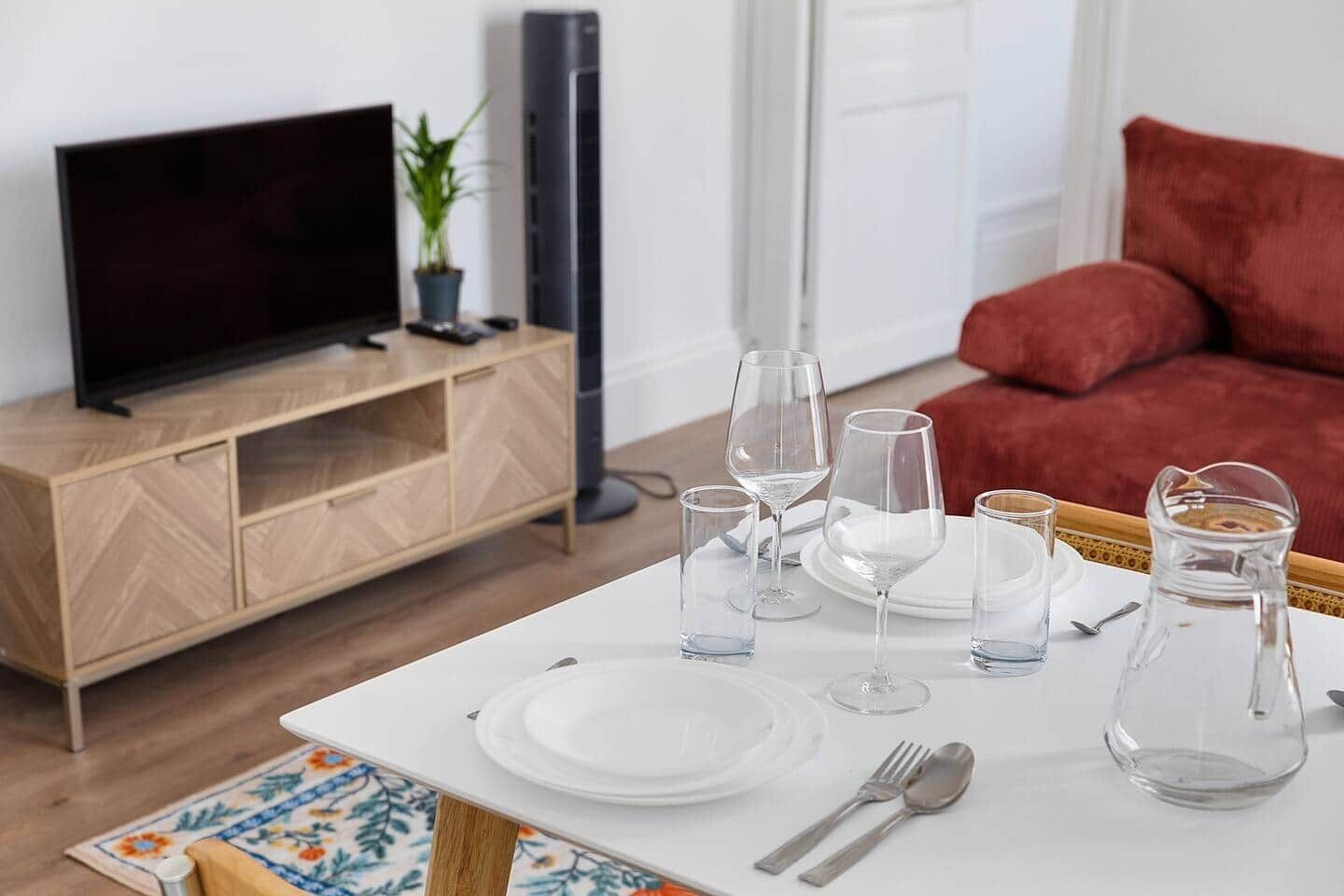 A dining table set with plates, glasses, and cutlery, a television on a wooden cabinet, and a red sofa in the background.