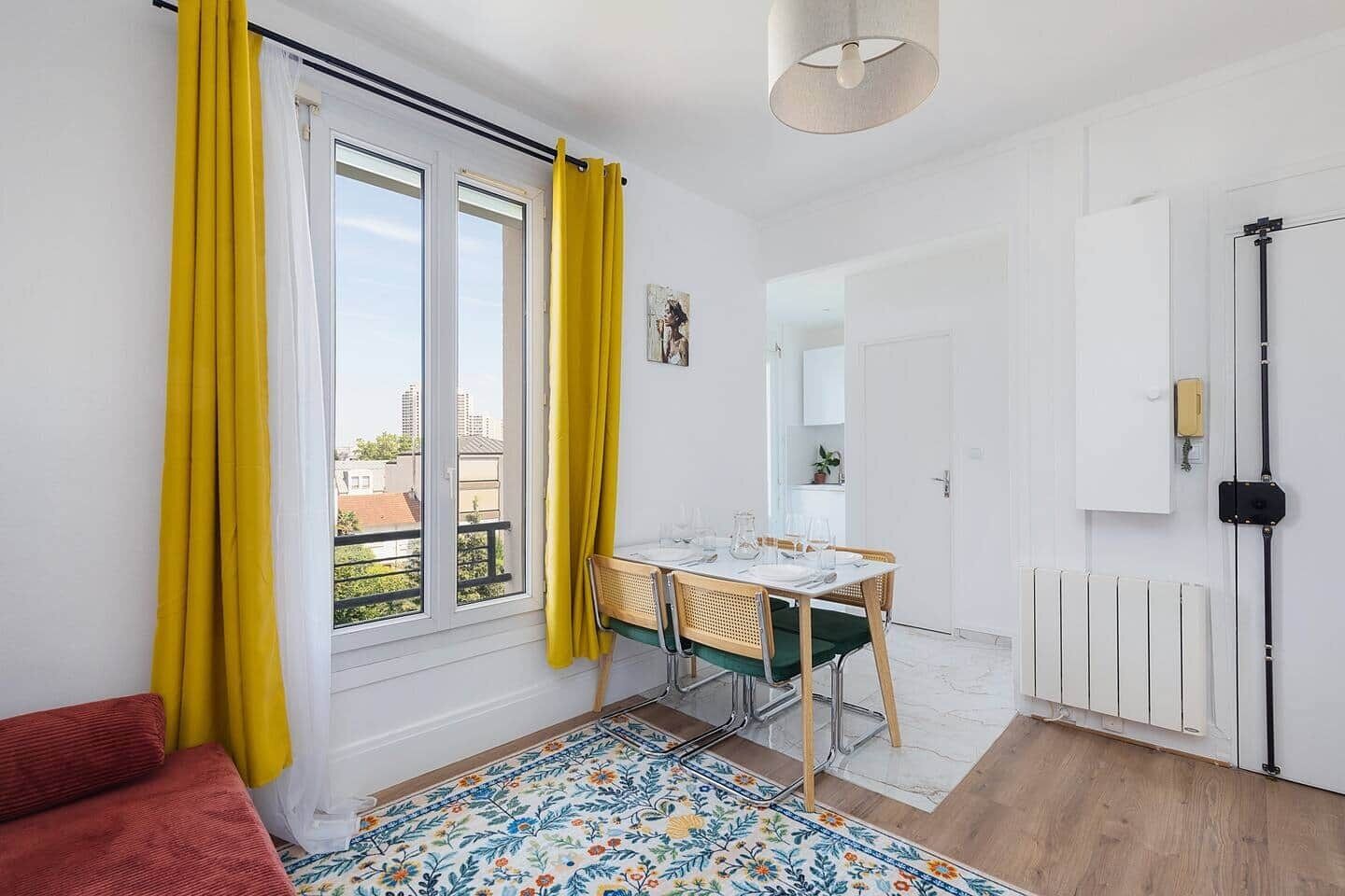 A room with a dining table, chairs, a red sofa, a patterned rug, a radiator, and a window with yellow curtains.