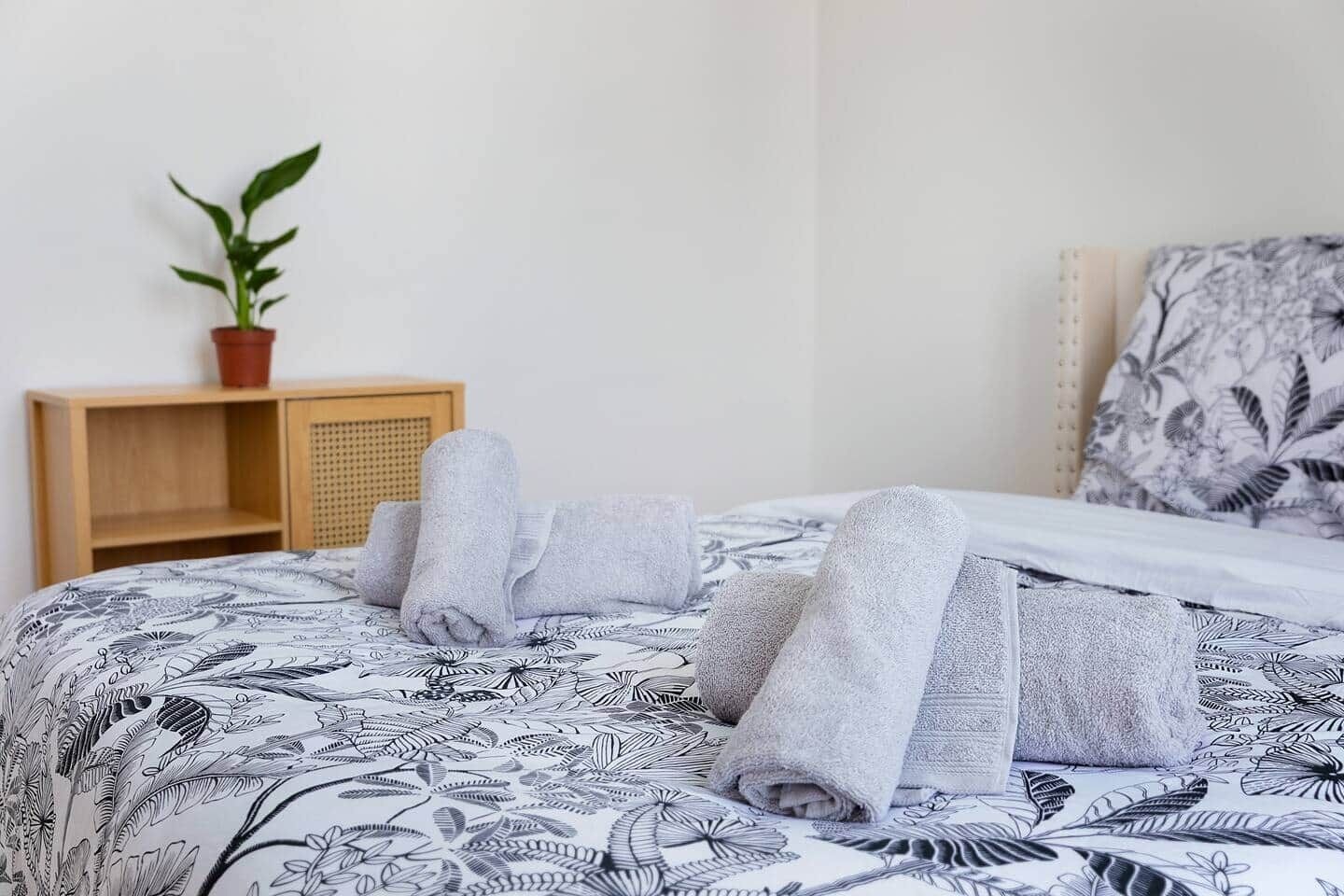 Un lit bien fait, recouvert d’une couvre-lit à motifs, une table de chevet en bois ornée d’une plante en pot et une tête de lit décorée d’un motif de feuilles.