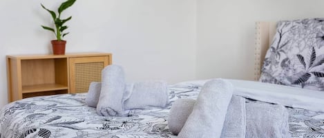 Un lit bien fait, recouvert d’une couvre-lit à motifs, une table de chevet en bois ornée d’une plante en pot et une tête de lit décorée d’un motif de feuilles.