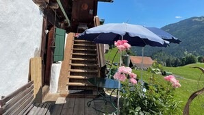 Eine Holztreppe führt zu einem Gebäude, im Vordergrund ein blauer Sonnenschirm und rosa Blumen.