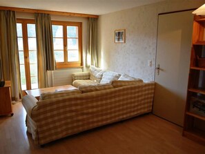 Ein Zimmer mit einem karierten Sofa, einem Holztisch, einem Fenster mit Vorhängen und einem Kleiderschrank.