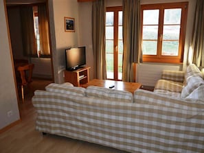 Un saló amb un sofà estampat de quadradets, una televisió sobre un moble de fusta, una zona de menjador amb una cadira i una finestra amb cortines.