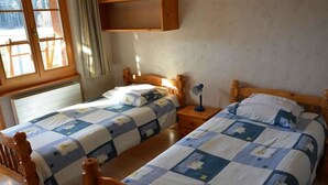 A room with two wooden beds, each with a quilted bedspread. There is a window with curtains, a wooden shelf above the beds, and a nightstand with a lamp. - Jacqueline 11 Appartement/Fewo, Dusche oder Bad, WC 6-Bettwohnung by Interhome (Gstaad)