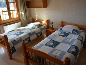 Ein Zimmer mit zwei Holzbett, jedes mit einer gesteppten Bettdecke. Es gibt ein Fenster mit Vorhängen, ein Holzregal über den Betten und einen Nachttisch mit einer Lampe.