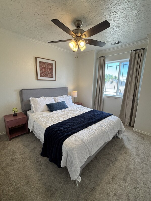 A bedroom with a bed, a nightstand, a ceiling fan, a window with curtains, and a framed picture on the wall. - Colorful Cottage — Spacious 4BR Family Home that Sleeps up to 12 (Twin Falls)