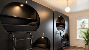 A room with bunk beds numbered 1 to 4, a wooden floor, and a window with a view of palm trees.