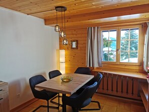 A dining area with a wooden table, black chairs, a hanging light fixture, and a window with a view of trees.