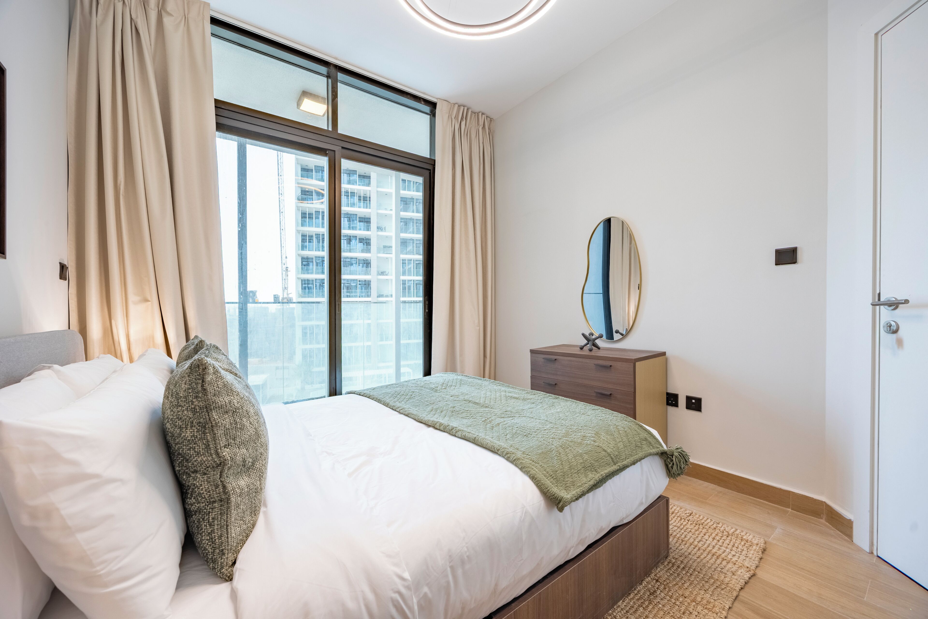 A bedroom with a large window, a bed with white bedding, a bedside table with a mirror, and a door.