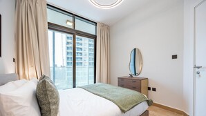 Une chambre avec une grande fenêtre, un lit avec une literie blanche, une table de chevet avec un miroir et une porte.