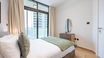 A bedroom with a large window, a bed with white bedding, a bedside table with a mirror, and a door.