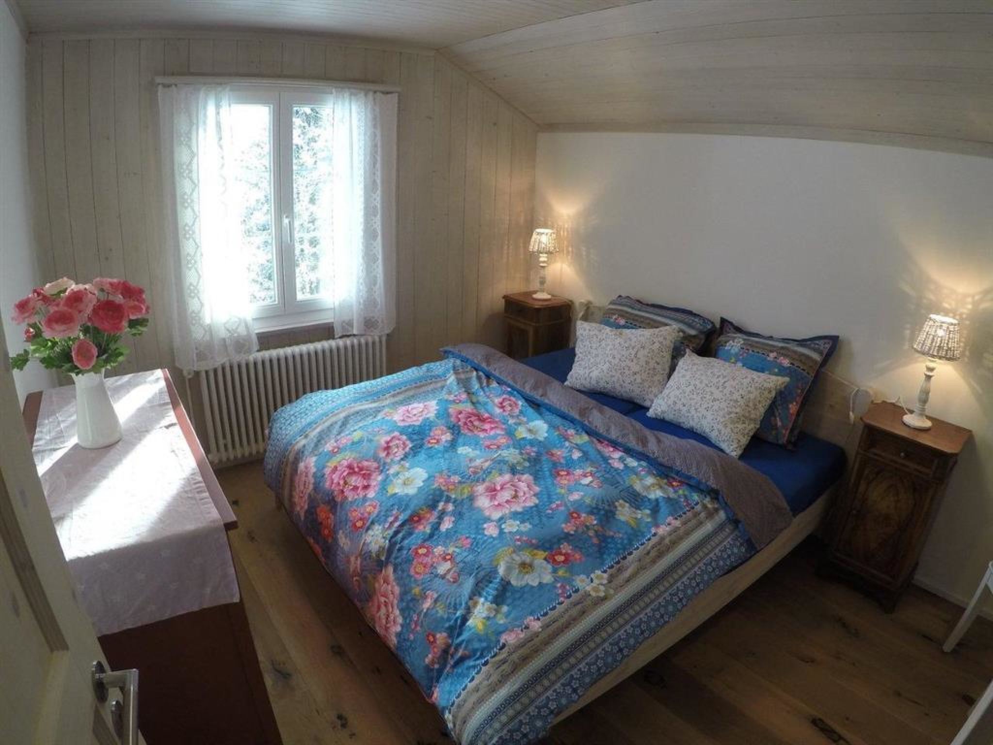 Une chambre à coucher avec une couvre-lit à motifs floraux, une fenêtre avec des rideaux, une lampe de chevet et un radiateur.