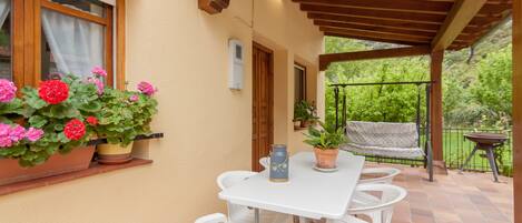 Un patio techado con una mesa y sillas, plantas en macetas y vistas al verdor.