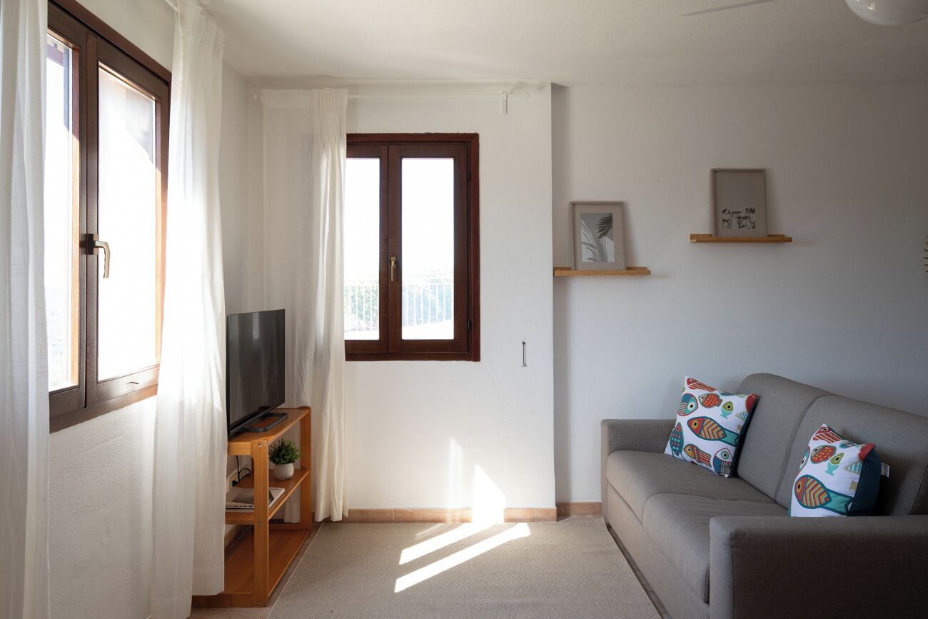 Una sala de estar con un televisor sobre un mueble de madera, un sofá con cojines estampados, una ventana con cortinas y estanterías de pared con cuadros enmarcados.