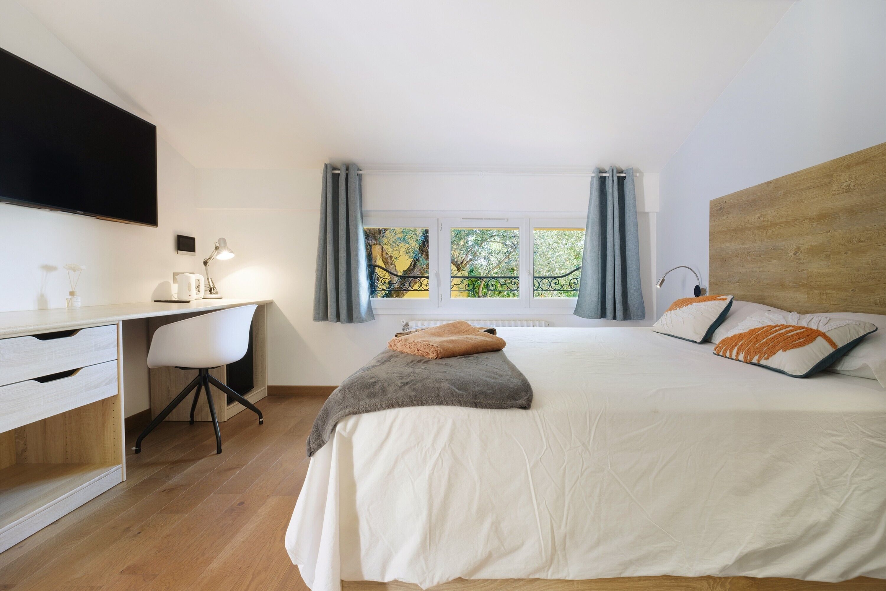 Ein Schlafzimmer mit einem Bett, einem Schreibtisch, einem Stuhl, einem an der Wand befestigten Fernseher und einem Fenster mit Vorhängen.