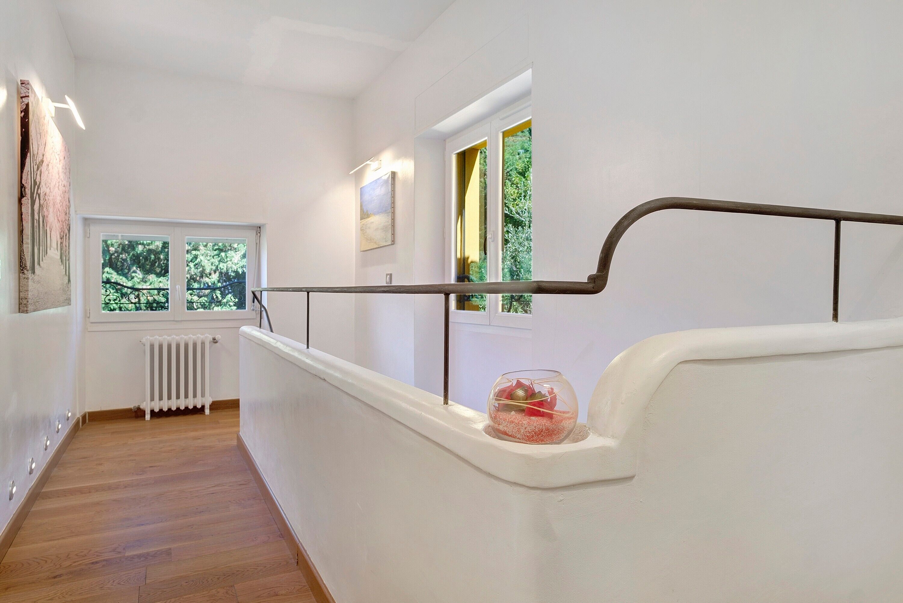 Un escalier avec une rampe blanche, un bol en verre contenant des objets rouges sur le palier, et un radiateur fixé au mur.