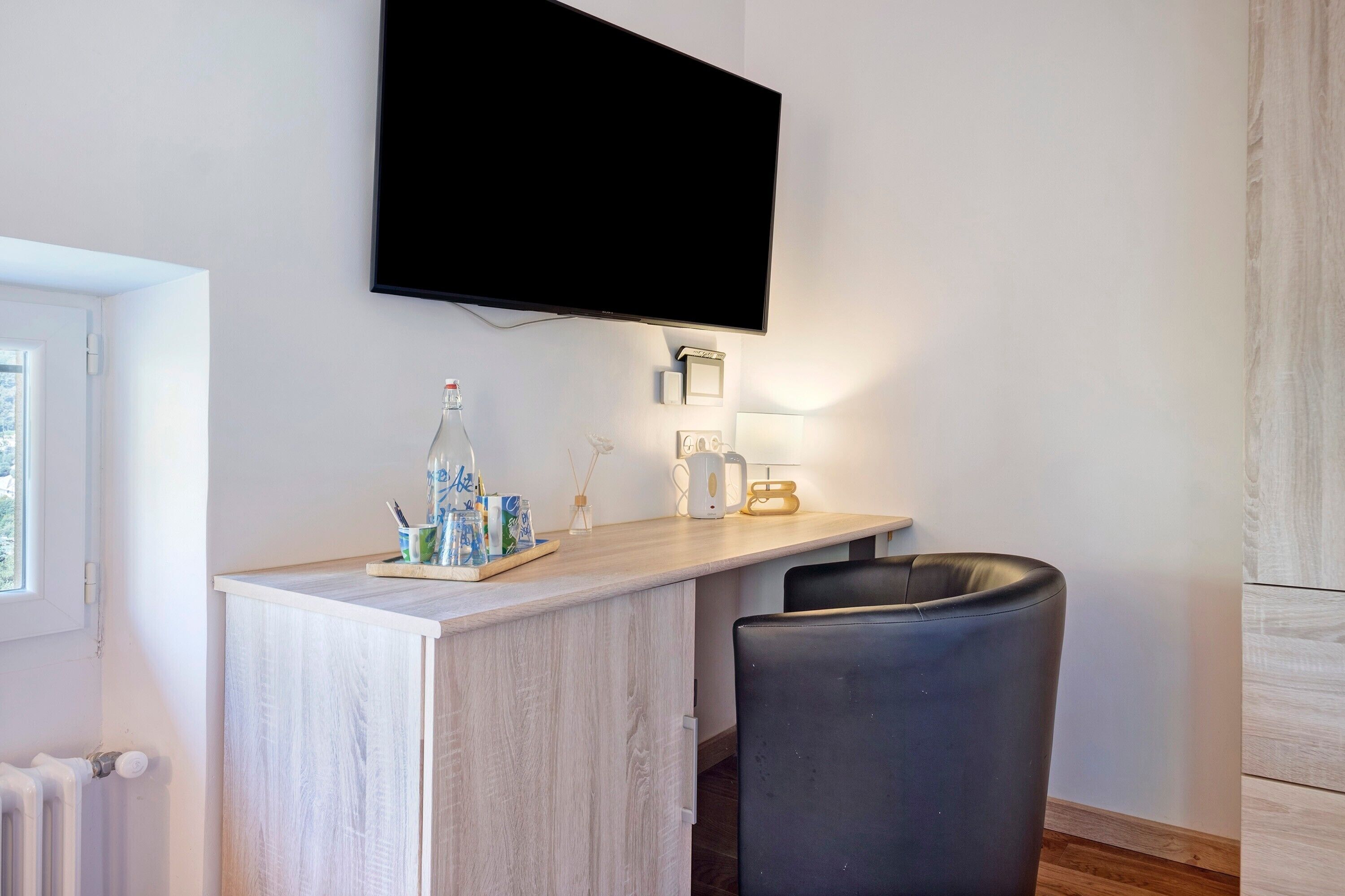 Une chambre moderne dotée d’un téléviseur à écran plat fixé au mur, d’un bureau en bois avec une chaise, d’un radiateur et d’une fenêtre.