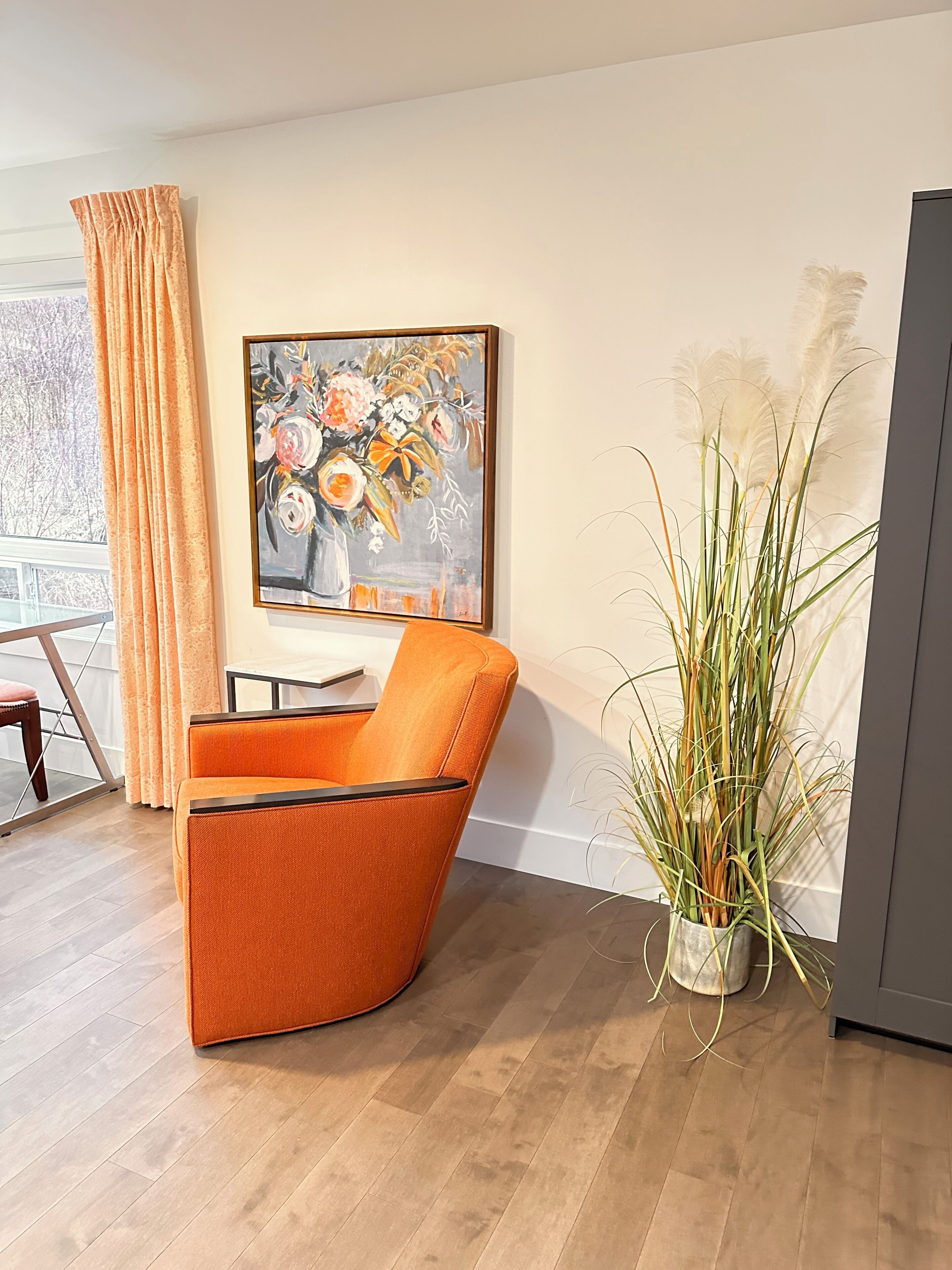 Une pièce avec un fauteuil orange aux lignes courbes, un tableau représentant des fleurs accroché au mur, une plante en pot et une vue par la fenêtre.
