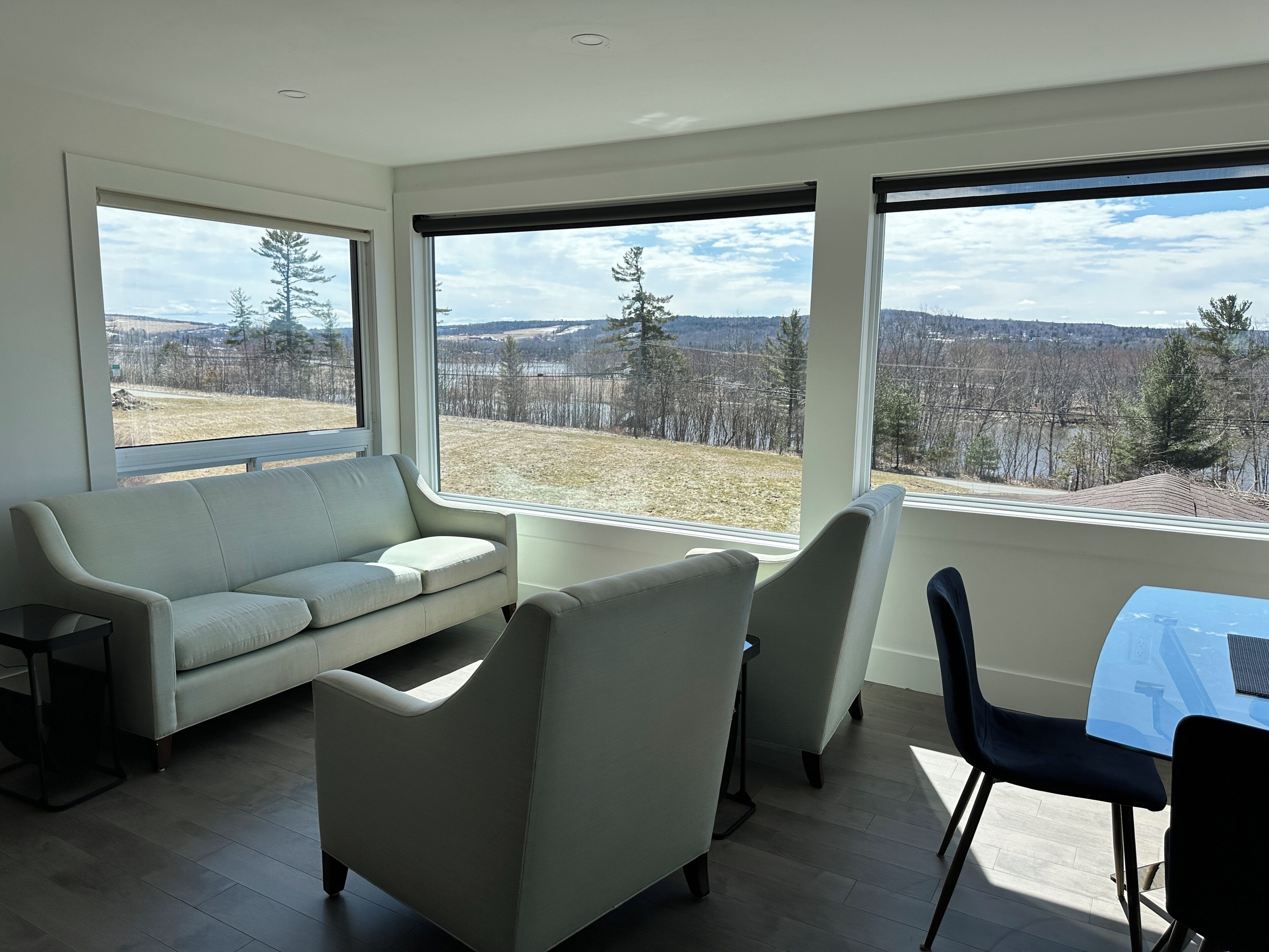 A modern living room with a sofa, chairs, a small table, and large windows offering a view of a rural landscape.