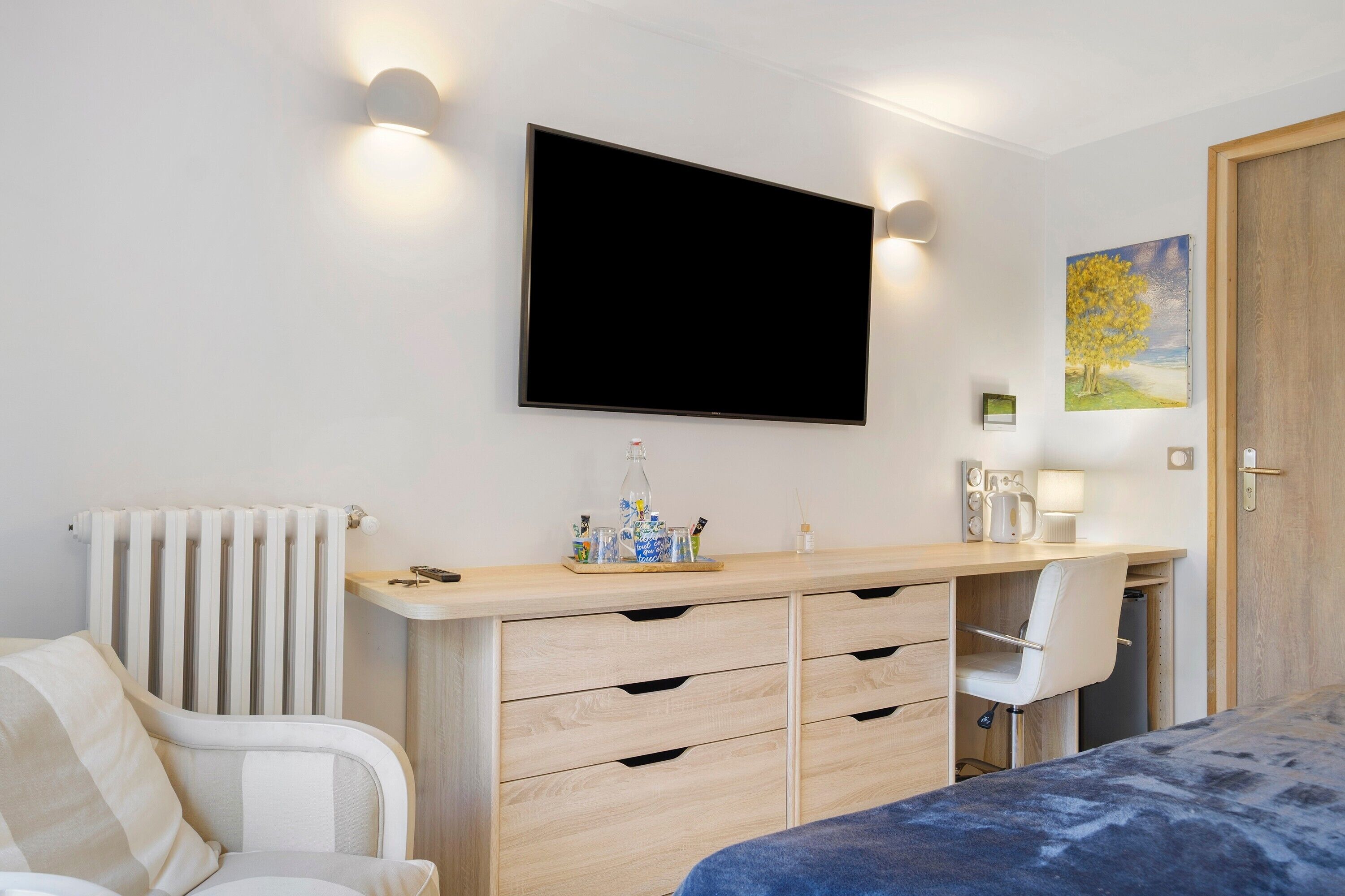 A modern bedroom with a bed, a desk, a chair, a radiator, a television, and a painting on the wall.