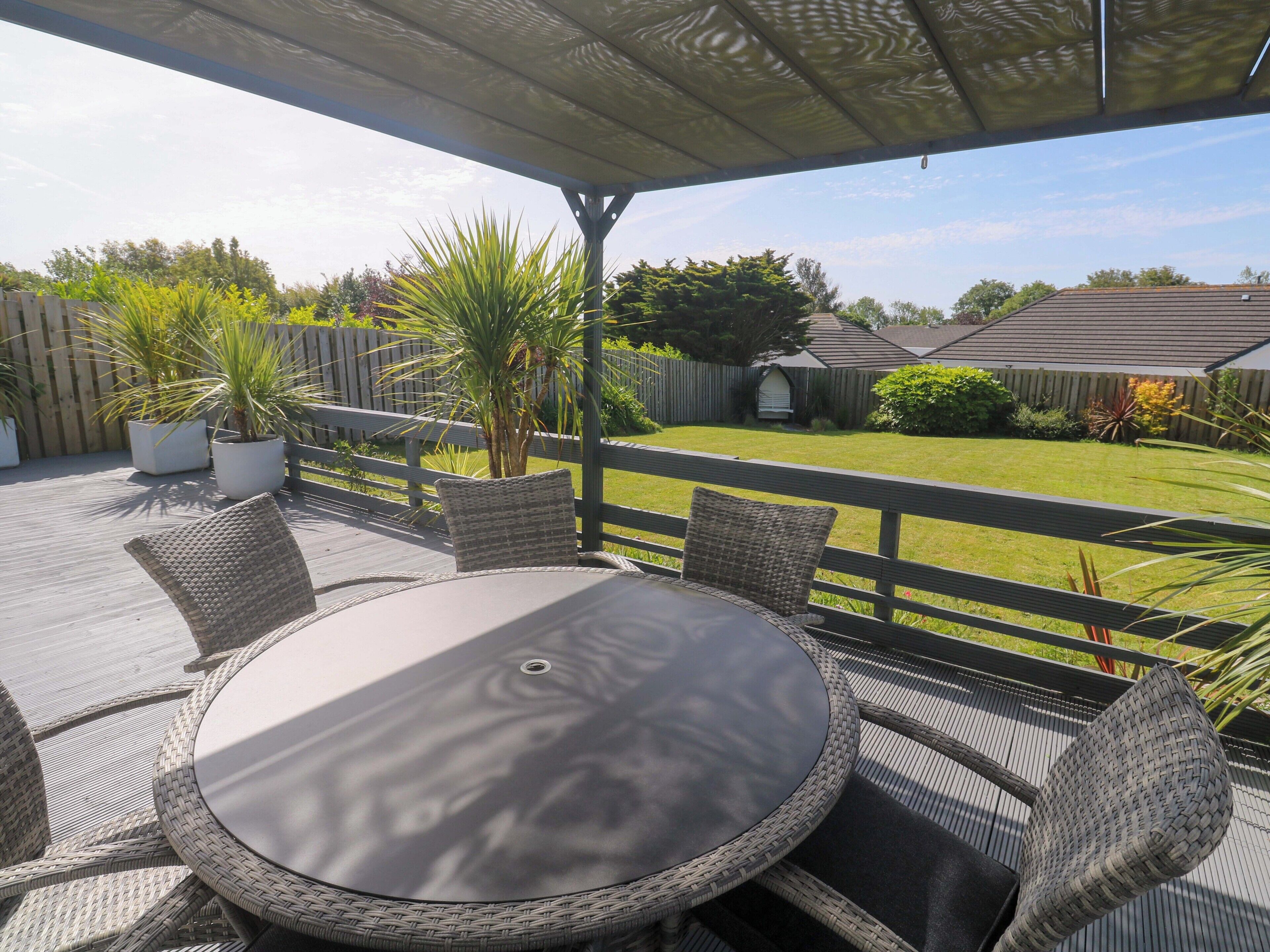 A covered patio with a round table and wicker chairs, overlooking a grassy yard and a wooden fence.
