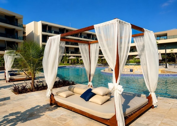Una cama junto a la piscina con dosel, una piscina y un edificio moderno al fondo.