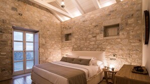 A stone-walled bedroom with a bed, a small table, and a view through a glass door.