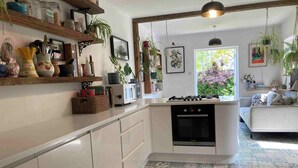 A kitchen with a central island, a stove, and a sink. There are shelves with jars and plants, and a window with a view of greenery. - Cambridge Center - Riverside - On a no-through road leading to the River Cam (Cambridge)