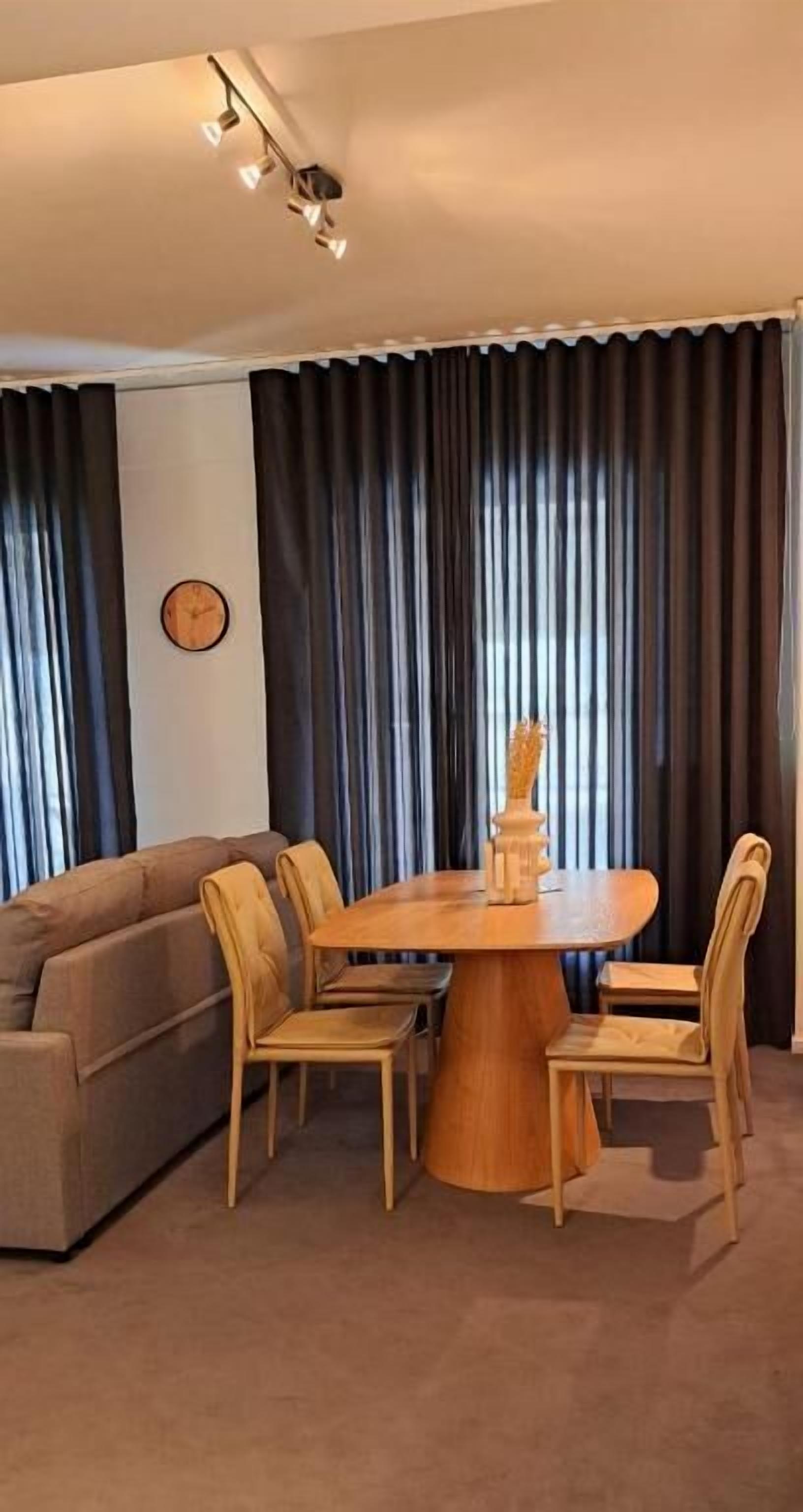 Un salon avec une table ronde en bois, quatre chaises, un canapé et une grande fenêtre ornée de rideaux foncés.
