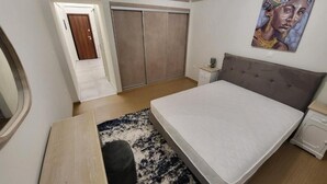 Un dormitorio con cama, mesita de noche, cómoda, armario, un cuadro en la pared y una puerta que da a otra habitación.