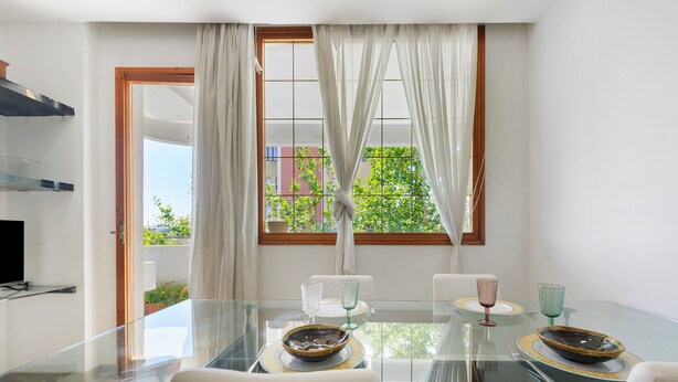 A modern dining area with a glass table, white chairs, and a view of greenery through a window.