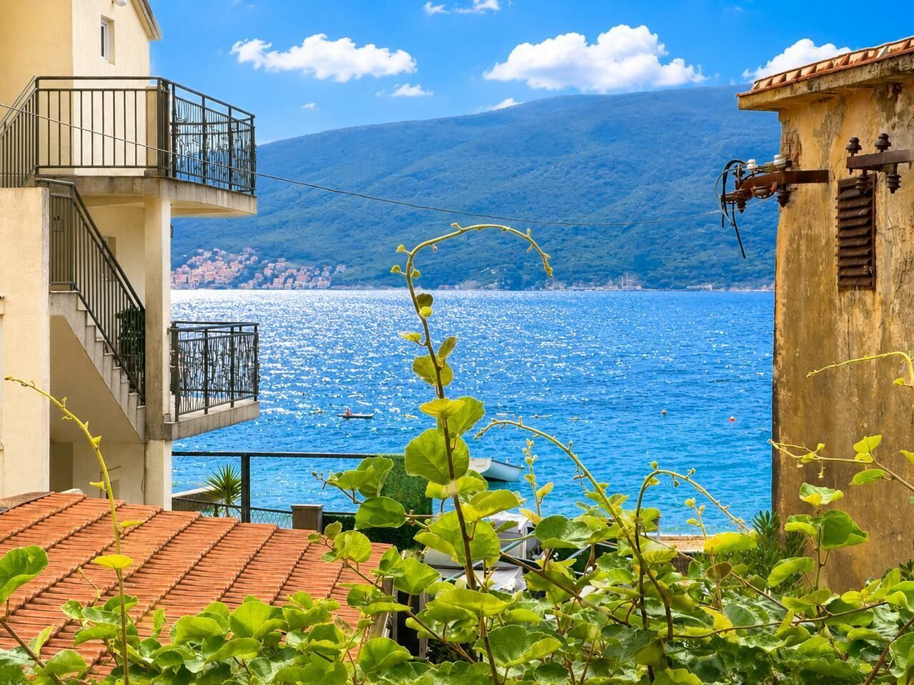 Ein Küstengebäude mit einem Balkon mit Blick auf das klare blaue Meer, einem gebirgigen Hintergrund und üppiger Vegetation im Vordergrund.