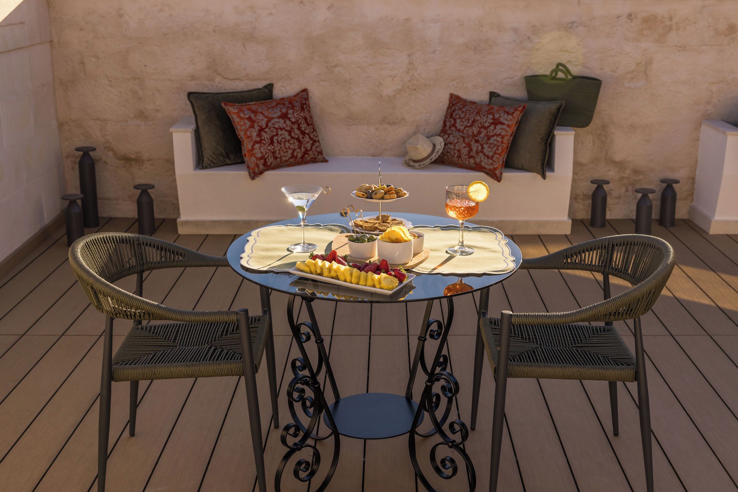 A round table with a white tablecloth, two wicker chairs, a sofa with patterned cushions, and a tray with food and drinks.