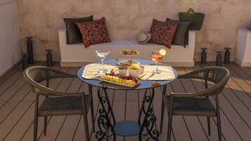 A round table with a white tablecloth, two wicker chairs, a sofa with patterned cushions, and a tray with food and drinks.