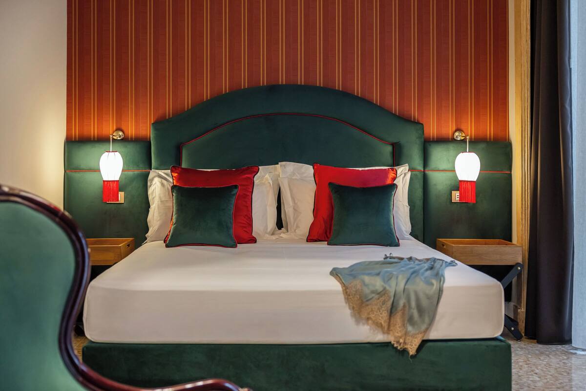Uma cama bem arrumada com cabeceira estofada verde, duas mesas de cabeceira com abajures e um criado-mudo de madeira.